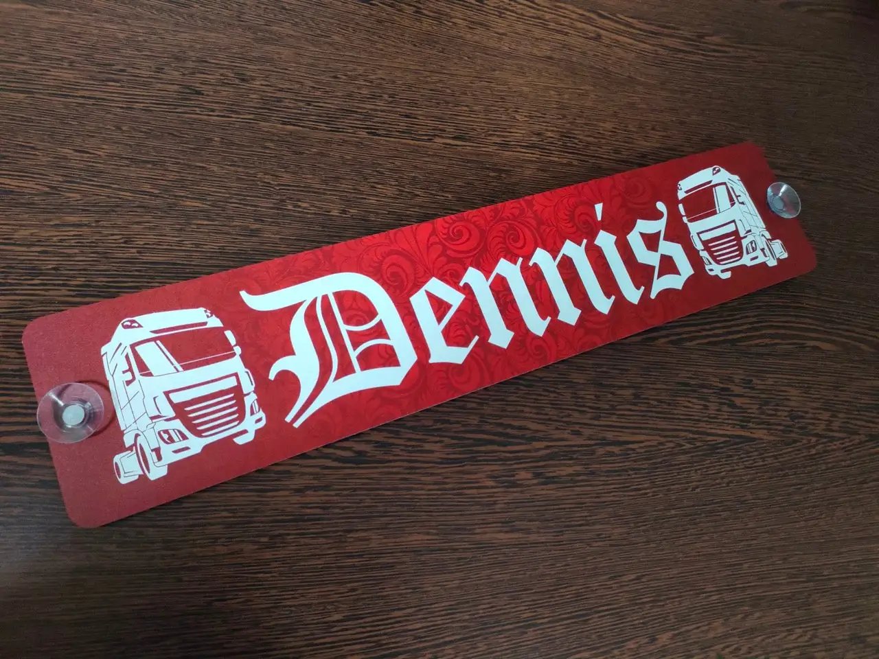 Custom LKW nameplate in red with white text "Dennis" and truck graphics, mounted with suction cups on a wooden surface.