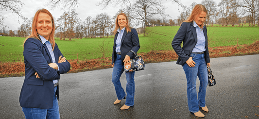 Woman in blue blazer and jeans walking on a rural road with fields in the background, holding a floral bag.