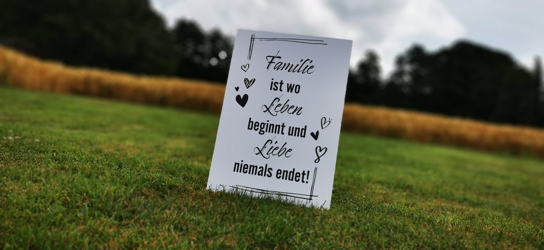 Decorative sign with German quote about family and love, placed on a grassy field with wheat and trees in the background.