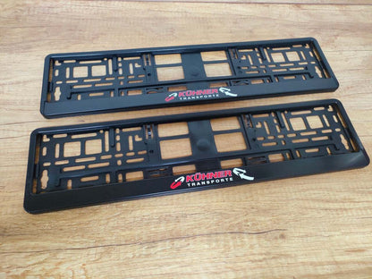Set of two black license plate holders with customizable print. Durable and weather-resistant materials showcased on a wooden surface.
