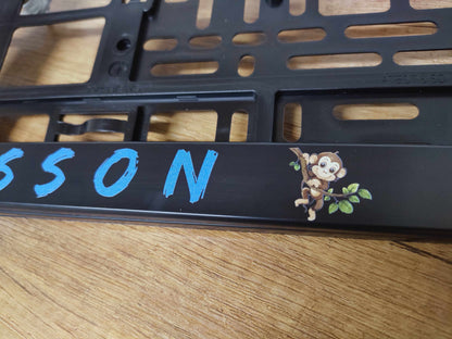 Black license plate holder with custom blue lettering and a cartoon monkey, made from polypropylene and polystyrol, UV-resistant.