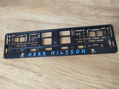 Custom black license plate holder with personalized text 'HERR NILSSON' in blue, featuring a sturdy polypropylene frame.