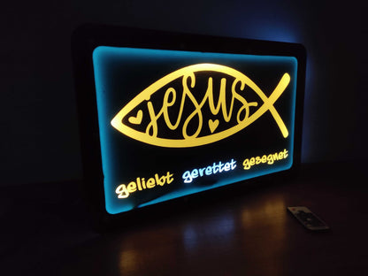 Illuminated LED sign with "Jesus" in a fish symbol, glowing in yellow and blue hues, featuring words "geliebt, gerettet, gesegnet".