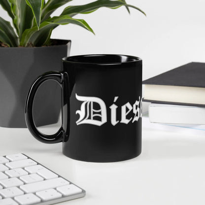 Schwarze glänzende Dieselknecht Tasse aus Keramik auf Schreibtisch, neben Buch und Pflanze