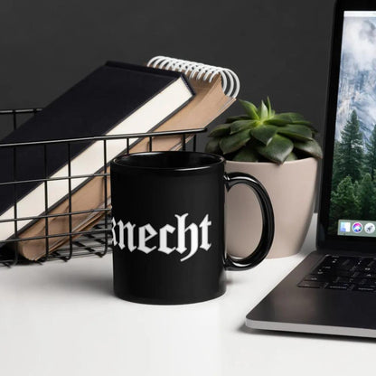 Schwarze glänzende Tasse mit 'Dieselknecht' Aufdruck, neben Laptop und Pflanze, ideal für stilvollen Kaffeegenuss.