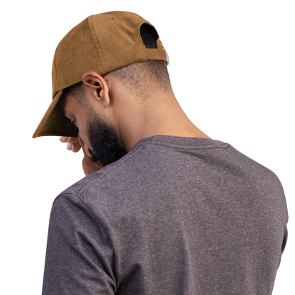 Person wearing a brown corduroy cap with cross embroidery, looking down, in a casual grey t-shirt, on a white background.