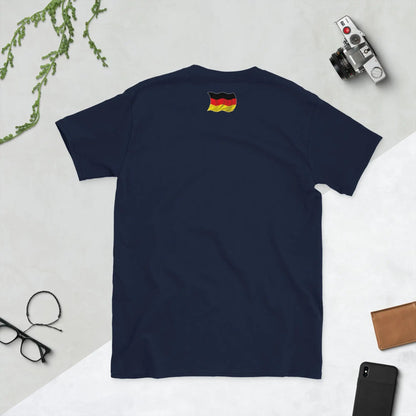 Rückansicht von dunkelblauem T-Shirt mit Deutschlandflagge, umgeben von Kamera, Pflanze und Handy auf hellem Hintergrund.