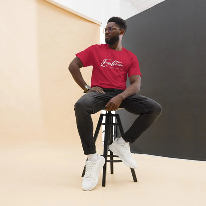 Man wearing a red T-shirt with 'Jesus Christ Experience' print, sitting on a stool against a minimalist backdrop.