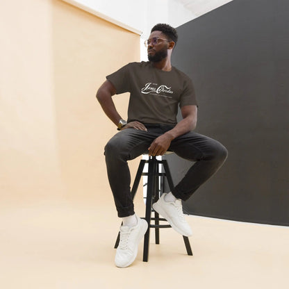 Man wearing 'Erlebe Jesus Christus' T-shirt sits on stool, showcasing faith-inspired apparel made of 100% cotton for casual style.