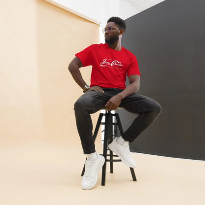 Man wearing a red T-shirt with 'Erlebe Jesus Christus und du wirst nie mehr Durst haben' text, sitting on a stool, showcasing faith-inspired fashion.