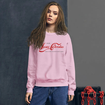 Woman wearing a pink UNI pullover with 'Erlebe Jesus Christus' design, standing against a dark wall.