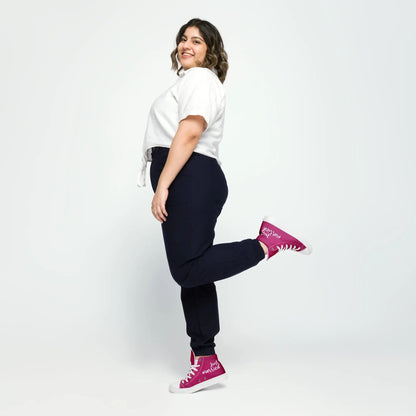 Frau in weißem Hemd und schwarzen Hosen, trägt "Just married" Sneaker in Pink, hebt ein Bein, posiert vor neutralem Hintergrund.