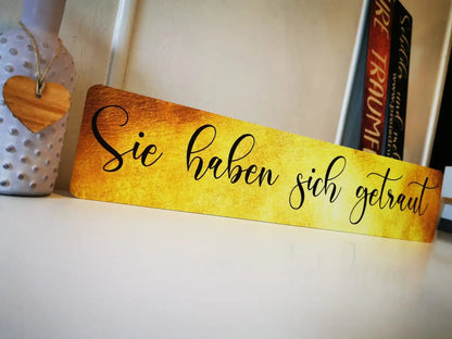 Image of a wedding sign with German text 'Sie haben sich getraut' on a golden background, placed on a shelf next to a decorative vase.