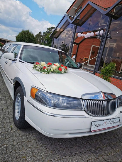 White wedding limousine with floral arrangement and customized license plate in front of a decorated venue.