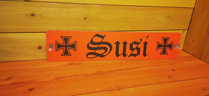 Orange personalized LKW nameplate with 'Susi' in black script, flanked by black crosses, on a wood background.