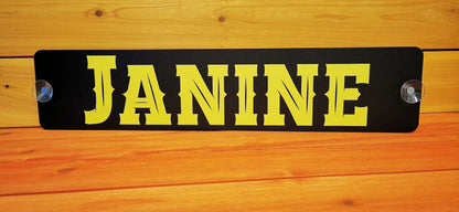 Black and yellow LKW windshield nameplate with "JANINE" displayed, mounted on wooden background.