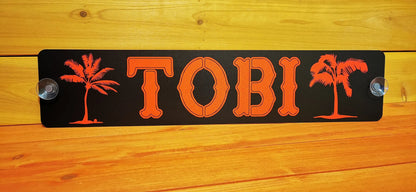 Custom LKW Namensschild with "TOBI" design, featuring palm tree motifs on a black background with attached suction cups for easy mounting.