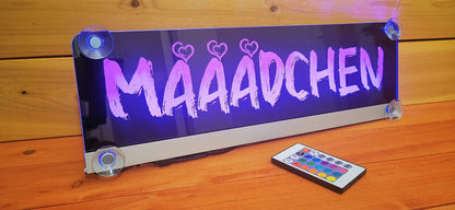 LED truck nameplate with suction cups, displaying 'Mädchen' in vibrant lighting, accompanied by a remote control on a wooden surface.