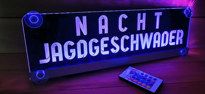 Illuminated LED name sign with suctions cups displaying 'Nacht Jagdgeschwader', remote control on a wooden surface.