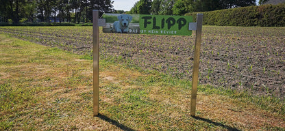 Wooden sign with a dog image and "FLIPP DAS IST MEIN REVIER" printed, standing on a grassy field edge near farmland.